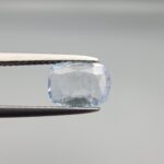 a premium, beautifully faceted 1.37-carat natural Blue Sapphire gemstone is held securely in metal tweezers against a clean, neutral grey background. The loose cushion-cut corundum gem displays a delicate icy light-blue hue with distinctive, beautiful internal silk and crystal inclusions that serve as a natural fingerprint, verifying its unheated earth-mined origin.