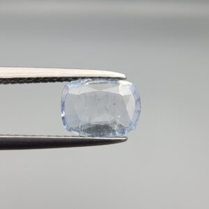 a premium, beautifully faceted 1.37-carat natural Blue Sapphire gemstone is held securely in metal tweezers against a clean, neutral grey background. The loose cushion-cut corundum gem displays a delicate icy light-blue hue with distinctive, beautiful internal silk and crystal inclusions that serve as a natural fingerprint, verifying its unheated earth-mined origin.