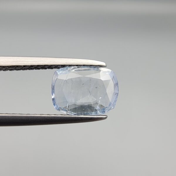 a premium, beautifully faceted 1.37-carat natural Blue Sapphire gemstone is held securely in metal tweezers against a clean, neutral grey background. The loose cushion-cut corundum gem displays a delicate icy light-blue hue with distinctive, beautiful internal silk and crystal inclusions that serve as a natural fingerprint, verifying its unheated earth-mined origin.
