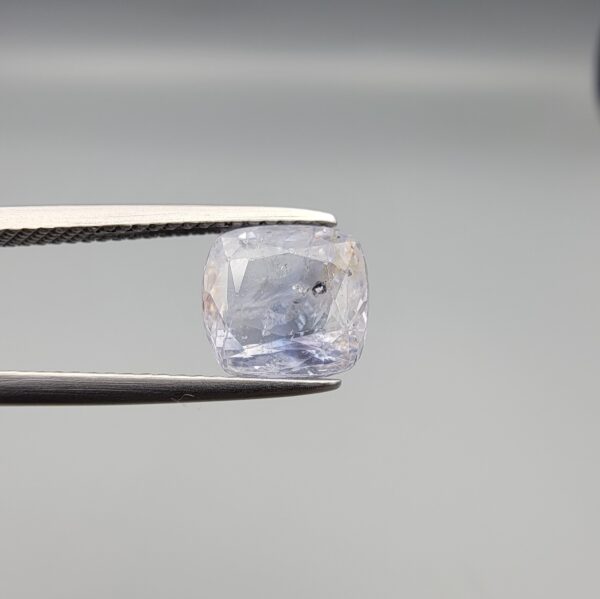 a premium, beautifully faceted 3.62-carat natural Blue Sapphire gemstone is held securely in metal tweezers against a clean, neutral grey background. The loose cushion-cut corundum gem displays a delicate icy light-blue color with a prominent natural internal cavity and fine crystal inclusions that serve as an indisputable signature of its unheated, earth-mined origin.