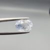 A premium, beautifully faceted 2.50-carat natural Blue Sapphire gemstone is held securely in metal tweezers against a clean, neutral grey background. The loose corundum gem showcases an expert oval cut with a well-proportioned table, displaying a serene, icy light-blue tone with fine, genuine internal crystal characteristics and silk inclusions that verify its rare, unheated earth-mined origin.