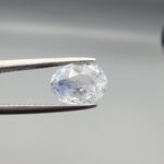A premium, beautifully faceted 2.50-carat natural Blue Sapphire gemstone is held securely in metal tweezers against a clean, neutral grey background. The loose corundum gem showcases an expert oval cut with a well-proportioned table, displaying a serene, icy light-blue tone with fine, genuine internal crystal characteristics and silk inclusions that verify its rare, unheated earth-mined origin.