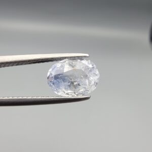 A premium, beautifully faceted 2.50-carat natural Blue Sapphire gemstone is held securely in metal tweezers against a clean, neutral grey background. The loose corundum gem showcases an expert oval cut with a well-proportioned table, displaying a serene, icy light-blue tone with fine, genuine internal crystal characteristics and silk inclusions that verify its rare, unheated earth-mined origin.