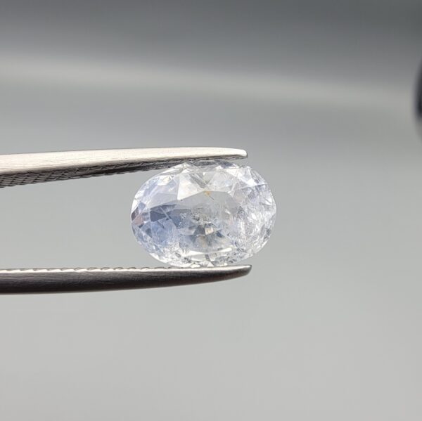 A premium, beautifully faceted 2.50-carat natural Blue Sapphire gemstone is held securely in metal tweezers against a clean, neutral grey background. The loose corundum gem showcases an expert oval cut with a well-proportioned table, displaying a serene, icy light-blue tone with fine, genuine internal crystal characteristics and silk inclusions that verify its rare, unheated earth-mined origin.