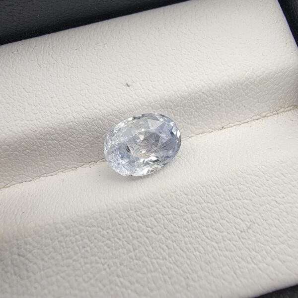 A premium, beautifully faceted 2.50-carat natural Blue Sapphire gemstone is held securely in metal tweezers against a clean, neutral grey background. The loose corundum gem showcases an expert oval cut with a well-proportioned table, displaying a serene, icy light-blue tone with fine, genuine internal crystal characteristics and silk inclusions that verify its rare, unheated earth-mined origin.