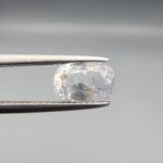 A premium, beautifully faceted 2.36-carat natural Blue Sapphire gemstone is held securely in metal tweezers against a clean, neutral grey background. The loose cushion-cut corundum gem displays a soft icy light-blue tone with fine internal silk and distinct golden-yellow mineral inclusions that serve as an indisputable signature of its unheated, earth-mined origin.