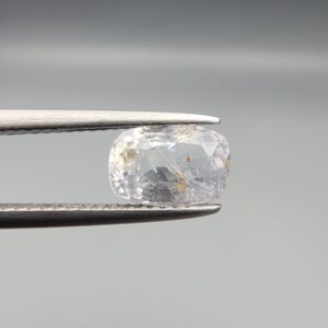 A premium, beautifully faceted 2.36-carat natural Blue Sapphire gemstone is held securely in metal tweezers against a clean, neutral grey background. The loose cushion-cut corundum gem displays a soft icy light-blue tone with fine internal silk and distinct golden-yellow mineral inclusions that serve as an indisputable signature of its unheated, earth-mined origin.