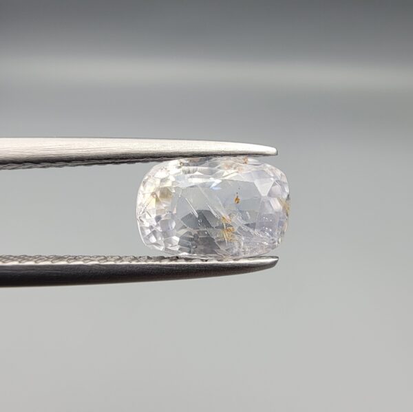 A premium, beautifully faceted 2.36-carat natural Blue Sapphire gemstone is held securely in metal tweezers against a clean, neutral grey background. The loose cushion-cut corundum gem displays a soft icy light-blue tone with fine internal silk and distinct golden-yellow mineral inclusions that serve as an indisputable signature of its unheated, earth-mined origin.