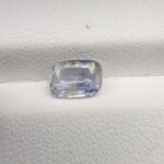 A premium, beautifully faceted 1.62-carat natural Blue Sapphire gemstone rests inside a white leatherette display box. The loose cushion-cut corundum gem displays a soft icy light-blue tone with fine internal silk and distinct golden-yellow mineral inclusions that serve as an indisputable signature of its unheated, earth-mined origin.