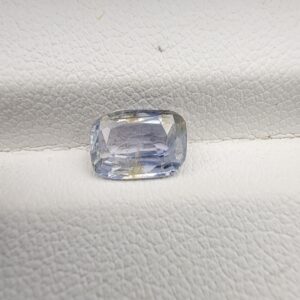 A premium, beautifully faceted 1.62-carat natural Blue Sapphire gemstone rests inside a white leatherette display box. The loose cushion-cut corundum gem displays a soft icy light-blue tone with fine internal silk and distinct golden-yellow mineral inclusions that serve as an indisputable signature of its unheated, earth-mined origin.