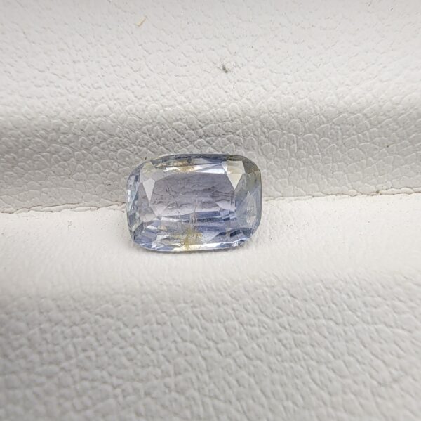 A premium, beautifully faceted 1.62-carat natural Blue Sapphire gemstone rests inside a white leatherette display box. The loose cushion-cut corundum gem displays a soft icy light-blue tone with fine internal silk and distinct golden-yellow mineral inclusions that serve as an indisputable signature of its unheated, earth-mined origin.