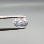 A premium, beautifully faceted 1.93-carat natural Blue Sapphire gemstone is held securely in metal tweezers against a clean, neutral grey background. The loose oval-cut corundum gem displays a soft icy light-blue tone with fine internal silk and distinct golden-yellow mineral inclusions that serve as an indisputable signature of its unheated, earth-mined origin.