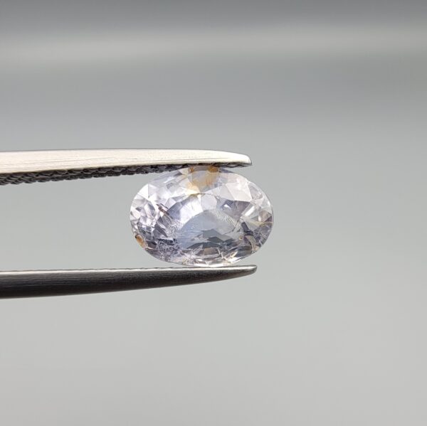 A premium, beautifully faceted 1.93-carat natural Blue Sapphire gemstone is held securely in metal tweezers against a clean, neutral grey background. The loose oval-cut corundum gem displays a soft icy light-blue tone with fine internal silk and distinct golden-yellow mineral inclusions that serve as an indisputable signature of its unheated, earth-mined origin.