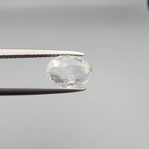 A premium, beautifully faceted 2.07-carat natural White Sapphire gemstone is held securely in metal tweezers against a clean, neutral grey background. The loose oval-cut corundum gem displays a completely colorless, diamond-like clarity with fine internal silk and natural crystal growth features that serve as an indisputable signature of its unheated, earth-mined origin.