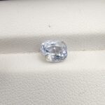 A premium, beautifully faceted 2.00-carat natural Blue Sapphire gemstone rests inside a white leatherette display box. The loose cushion-cut corundum gem displays a soft icy light-blue tone with fine internal silk and distinct golden-yellow mineral inclusions that serve as an indisputable signature of its unheated, earth-mined origin.
