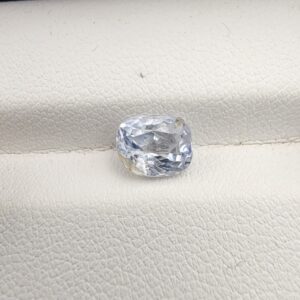 A premium, beautifully faceted 2.00-carat natural Blue Sapphire gemstone rests inside a white leatherette display box. The loose cushion-cut corundum gem displays a soft icy light-blue tone with fine internal silk and distinct golden-yellow mineral inclusions that serve as an indisputable signature of its unheated, earth-mined origin.