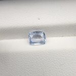 A premium, beautifully faceted 1.27-carat natural Blue Sapphire gemstone rests inside a white leatherette display box. The loose cushion-cut corundum gem displays a serene, icy light-blue tone with fine internal silk and natural micro-crystal inclusions that serve as an indisputable signature of its unheated, earth-mined origin.