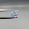 A premium, beautifully faceted 0.78-carat natural Blue Sapphire gemstone is held securely in metal tweezers against a clean, neutral grey background. The loose round-oval mixed cut corundum gem displays a serene light-blue hue with delicate internal silk and genuine crystal growth traits that serve as an indisputable signature of its unheated, earth-mined origin.