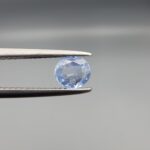 A premium, beautifully faceted 0.78-carat natural Blue Sapphire gemstone is held securely in metal tweezers against a clean, neutral grey background. The loose round-oval mixed cut corundum gem displays a serene light-blue hue with delicate internal silk and genuine crystal growth traits that serve as an indisputable signature of its unheated, earth-mined origin.