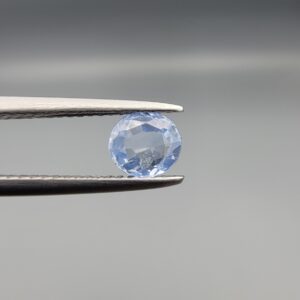 A premium, beautifully faceted 0.78-carat natural Blue Sapphire gemstone is held securely in metal tweezers against a clean, neutral grey background. The loose round-oval mixed cut corundum gem displays a serene light-blue hue with delicate internal silk and genuine crystal growth traits that serve as an indisputable signature of its unheated, earth-mined origin.