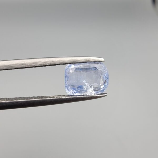 A premium, beautifully faceted 1.93-carat natural Blue Sapphire gemstone is held securely in metal tweezers against a clean, neutral grey background. The loose cushion-cut corundum gem displays a soft icy light-blue tone with fine internal silk and natural mineral inclusions that serve as an indisputable signature of its unheated, earth-mined origin.