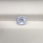 A premium, beautifully faceted 1.36-carat natural Blue Sapphire gemstone rests inside a white leatherette display box. The loose cushion-cut corundum gem displays a soft, icy light-blue tone with fine internal silk and genuine crystal growth characteristics that serve as an indisputable signature of its unheated, earth-mined origin.