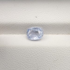 A premium, beautifully faceted 1.36-carat natural Blue Sapphire gemstone rests inside a white leatherette display box. The loose cushion-cut corundum gem displays a soft, icy light-blue tone with fine internal silk and genuine crystal growth characteristics that serve as an indisputable signature of its unheated, earth-mined origin.