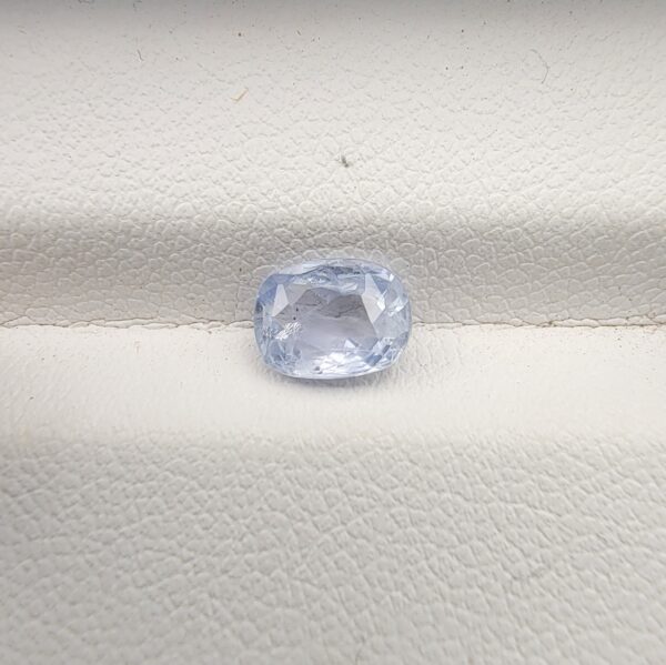 A premium, beautifully faceted 1.36-carat natural Blue Sapphire gemstone rests inside a white leatherette display box. The loose cushion-cut corundum gem displays a soft, icy light-blue tone with fine internal silk and genuine crystal growth characteristics that serve as an indisputable signature of its unheated, earth-mined origin.