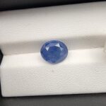 A premium, beautifully faceted 4.85-carat natural Blue Sapphire gemstone is held securely in metal tweezers against a clean, neutral grey background. The loose oval-cut corundum gem displays a vibrant, saturated blue tone with delicate internal silk and natural crystal inclusions that serve as an indisputable signature of its unheated, earth-mined origin.