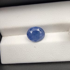 A premium, beautifully faceted 4.85-carat natural Blue Sapphire gemstone is held securely in metal tweezers against a clean, neutral grey background. The loose oval-cut corundum gem displays a vibrant, saturated blue tone with delicate internal silk and natural crystal inclusions that serve as an indisputable signature of its unheated, earth-mined origin.