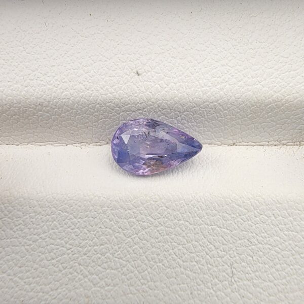 A premium, beautifully faceted 1.85-carat natural Purplish Blue Sapphire gemstone rests inside a white leatherette display box. The loose pear-cut corundum gem displays a unique violet-blue body tone with soft yellow mineral inclusions that serve as an indisputable signature of its unheated, earth-mined origin.