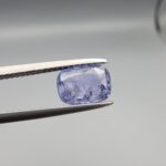 A premium, beautifully faceted 1.63-carat natural Violet Blue Sapphire gemstone is held securely in metal tweezers against a clean, neutral grey background. The loose cushion-cut corundum gem displays a serene violet-blue body tone with intricate internal mineral inclusions and fine silk that serve as an indisputable signature of its unheated, earth-mined origin.