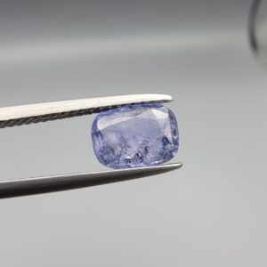 A premium, beautifully faceted 1.63-carat natural Violet Blue Sapphire gemstone is held securely in metal tweezers against a clean, neutral grey background. The loose cushion-cut corundum gem displays a serene violet-blue body tone with intricate internal mineral inclusions and fine silk that serve as an indisputable signature of its unheated, earth-mined origin.