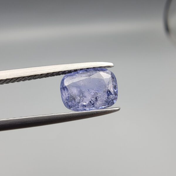 A premium, beautifully faceted 1.63-carat natural Violet Blue Sapphire gemstone is held securely in metal tweezers against a clean, neutral grey background. The loose cushion-cut corundum gem displays a serene violet-blue body tone with intricate internal mineral inclusions and fine silk that serve as an indisputable signature of its unheated, earth-mined origin.