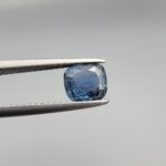 A premium, beautifully faceted 1.42-carat natural Deep Blue Sapphire gemstone is held securely in metal tweezers against a clean, neutral grey background. The loose cushion-cut corundum gem displays a rich, intense deep-blue hue with fine internal silk and genuine crystal growth characteristics that serve as an indisputable signature of its unheated, earth-mined origin.