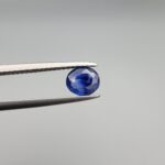 A premium, beautifully faceted 0.86-carat natural Blue Sapphire gemstone is held securely in metal tweezers against a clean, neutral grey background. The loose oval-cut corundum gem displays an intense, nearly royal blue hue with striking internal silk banding and natural growth features that serve as an indisputable signature of its unheated, earth-mined origin.