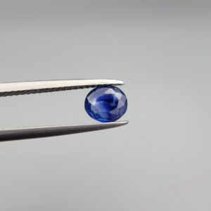A premium, beautifully faceted 0.86-carat natural Blue Sapphire gemstone is held securely in metal tweezers against a clean, neutral grey background. The loose oval-cut corundum gem displays an intense, nearly royal blue hue with striking internal silk banding and natural growth features that serve as an indisputable signature of its unheated, earth-mined origin.