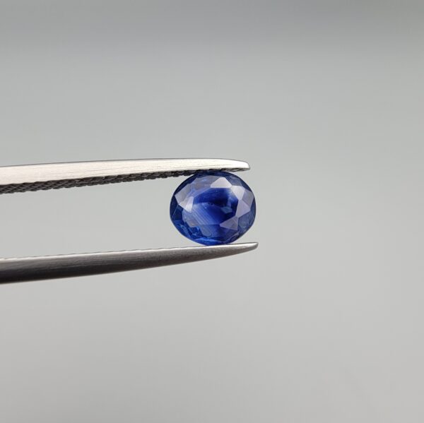 A premium, beautifully faceted 0.86-carat natural Blue Sapphire gemstone is held securely in metal tweezers against a clean, neutral grey background. The loose oval-cut corundum gem displays an intense, nearly royal blue hue with striking internal silk banding and natural growth features that serve as an indisputable signature of its unheated, earth-mined origin.