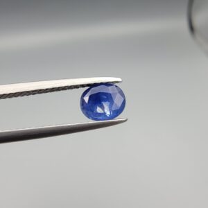A premium, beautifully faceted 0.96-carat natural Deep Blue Sapphire gemstone is held securely in metal tweezers against a clean, neutral grey background. The loose oval-cut corundum gem displays a rich, velvety deep-blue hue with striking internal silk reflection and natural growth features that serve as an indisputable signature of its unheated, earth-mined origin.