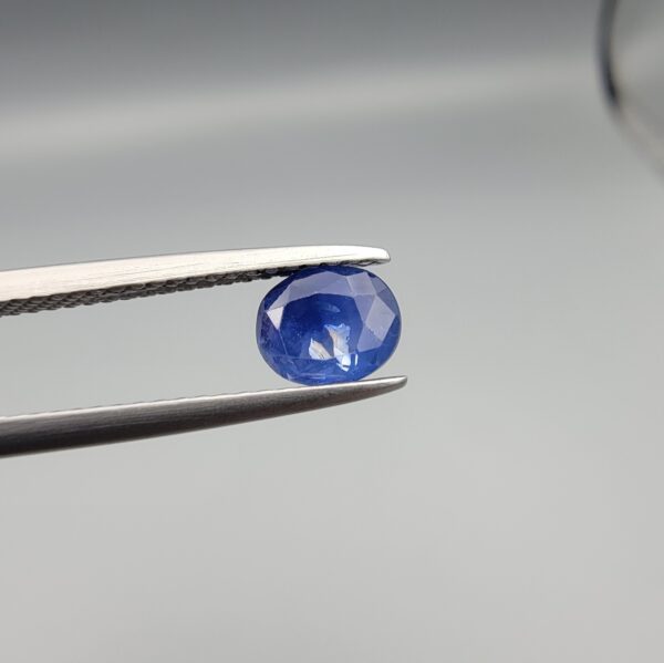A premium, beautifully faceted 0.96-carat natural Deep Blue Sapphire gemstone is held securely in metal tweezers against a clean, neutral grey background. The loose oval-cut corundum gem displays a rich, velvety deep-blue hue with striking internal silk reflection and natural growth features that serve as an indisputable signature of its unheated, earth-mined origin.