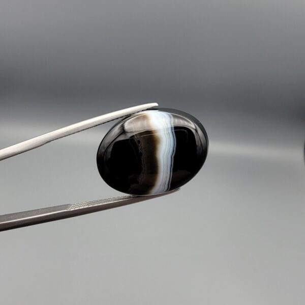 "Natural Black Banded Sulemani Aqeeq Agate oval cabochon gemstone 15.08 carat with vertical white and brown side band held in tweezers"