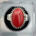 ​A premium men's luxury ring crafted from high-polish sterling silver, showcasing a massive 18.10-carat natural Japanese Red Coral (Marjan) cabochon as the central focal point. The vibrant red oval coral is securely held by a decorative multi-prong crown bezel setting on a thick, durable silver band, beautifully presented inside a professional display cushion case.