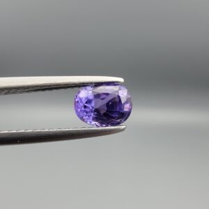 A premium, beautifully faceted 0.98-carat natural Purple Sapphire gemstone is held securely in metal tweezers against a clean, neutral grey background. The loose oval-cut corundum gem displays a vibrant, saturated purple hue, serving as an indisputable signature of its rare unheated, earth-mined origin.