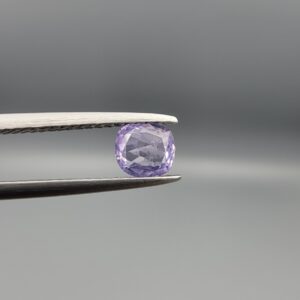 ​A premium, beautifully faceted 0.69-carat natural Purple Sapphire gemstone is held securely in metal tweezers against a clean, neutral grey background. The loose cushion-cut corundum gem displays a soft lavender-purple hue, serving as an indisputable signature of its rare unheated, earth-mined origin.