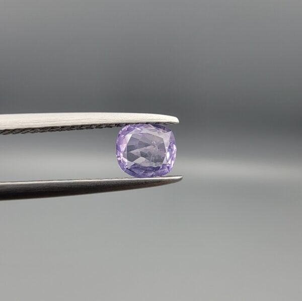 ​A premium, beautifully faceted 0.69-carat natural Purple Sapphire gemstone is held securely in metal tweezers against a clean, neutral grey background. The loose cushion-cut corundum gem displays a soft lavender-purple hue, serving as an indisputable signature of its rare unheated, earth-mined origin.