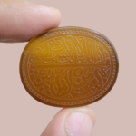A large 76.14-carat natural honey-amber Iranian chalcedony agate cabochon held between fingers, featuring intricate Arabic calligraphy engraving of Ayat-e-Karima (Dua-e-Younus) within an oval beaded border.
