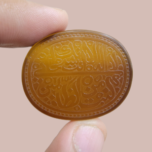 A large 76.14-carat natural honey-amber Iranian chalcedony agate cabochon held between fingers, featuring intricate Arabic calligraphy engraving of Ayat-e-Karima (Dua-e-Younus) within an oval beaded border.