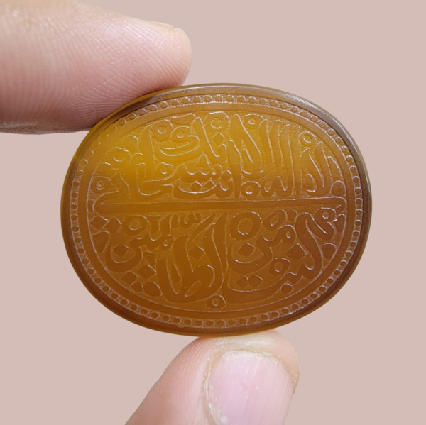 A large 76.14-carat natural honey-amber Iranian chalcedony agate cabochon held between fingers, featuring intricate Arabic calligraphy engraving of Ayat-e-Karima (Dua-e-Younus) within an oval beaded border.