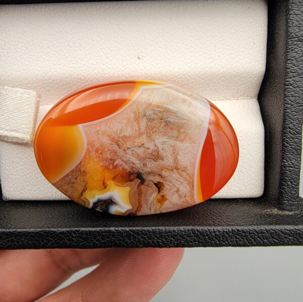 "Natural Carnelian Agate gemstone 85.42 carat oval cabochon from Yemen displayed in jewelry box showing red, orange, yellow, white, and brown natural color zones"
