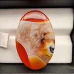 "Natural Carnelian Agate gemstone 85.42 carat oval cabochon from Yemen displayed in jewelry box showing red, orange, yellow, white, and brown natural color zones"